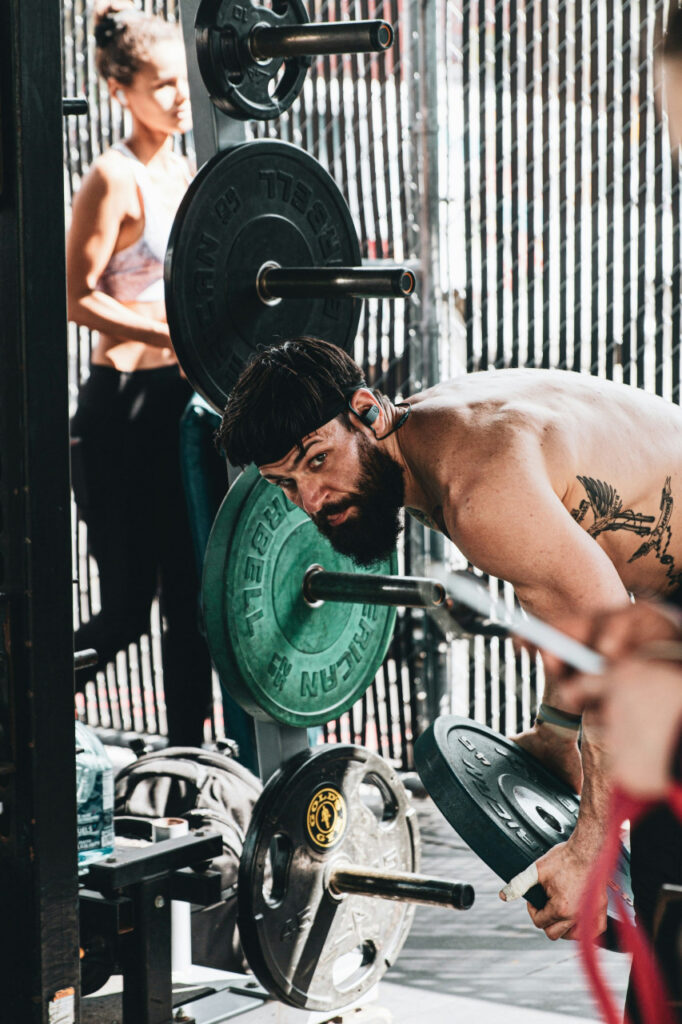 topless-man-carrying-barbell-in-front-of-woman-in-black-tank-top-XCkc8-_s7FA